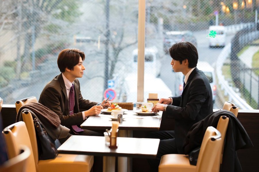os dois personagens principais juntos, sentados numa mesa tomando café e comendo doces enquanto sorriem sutilmente 